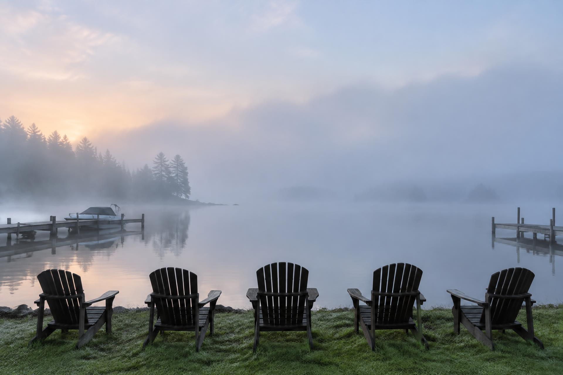 Five Adirondack chairs facing a still, misty lake at dawn